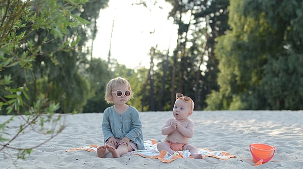 Two toddlers sit next to each other on a beach wearing orange sunglasses with beach toys nearby.